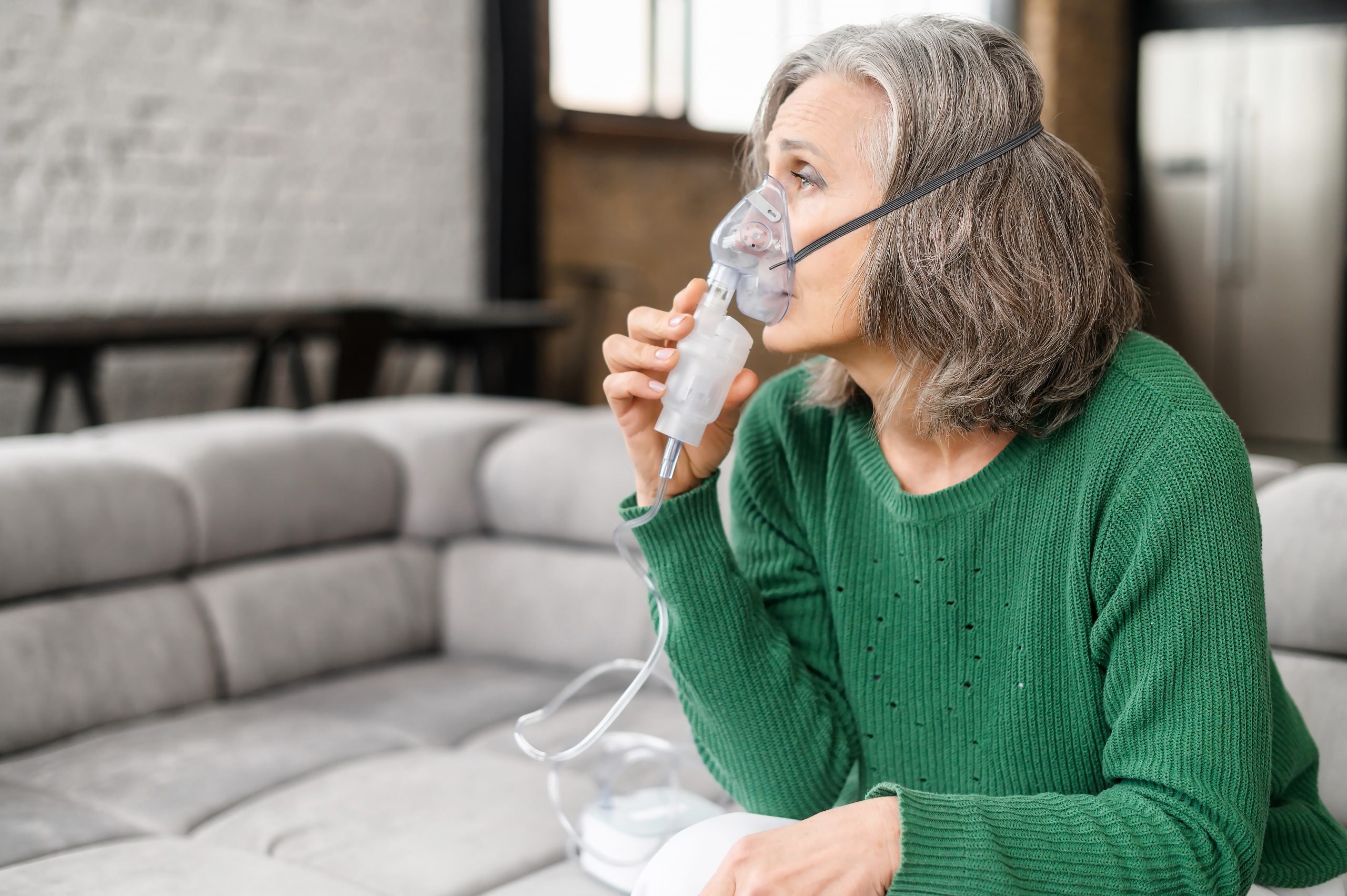 Upset senior woman feeling unwell, has shortness of breath, asthma, using oxygen mask sitting on the couch at home. Sick mature woman makes inhalation with a nebulizer at home, treating herself