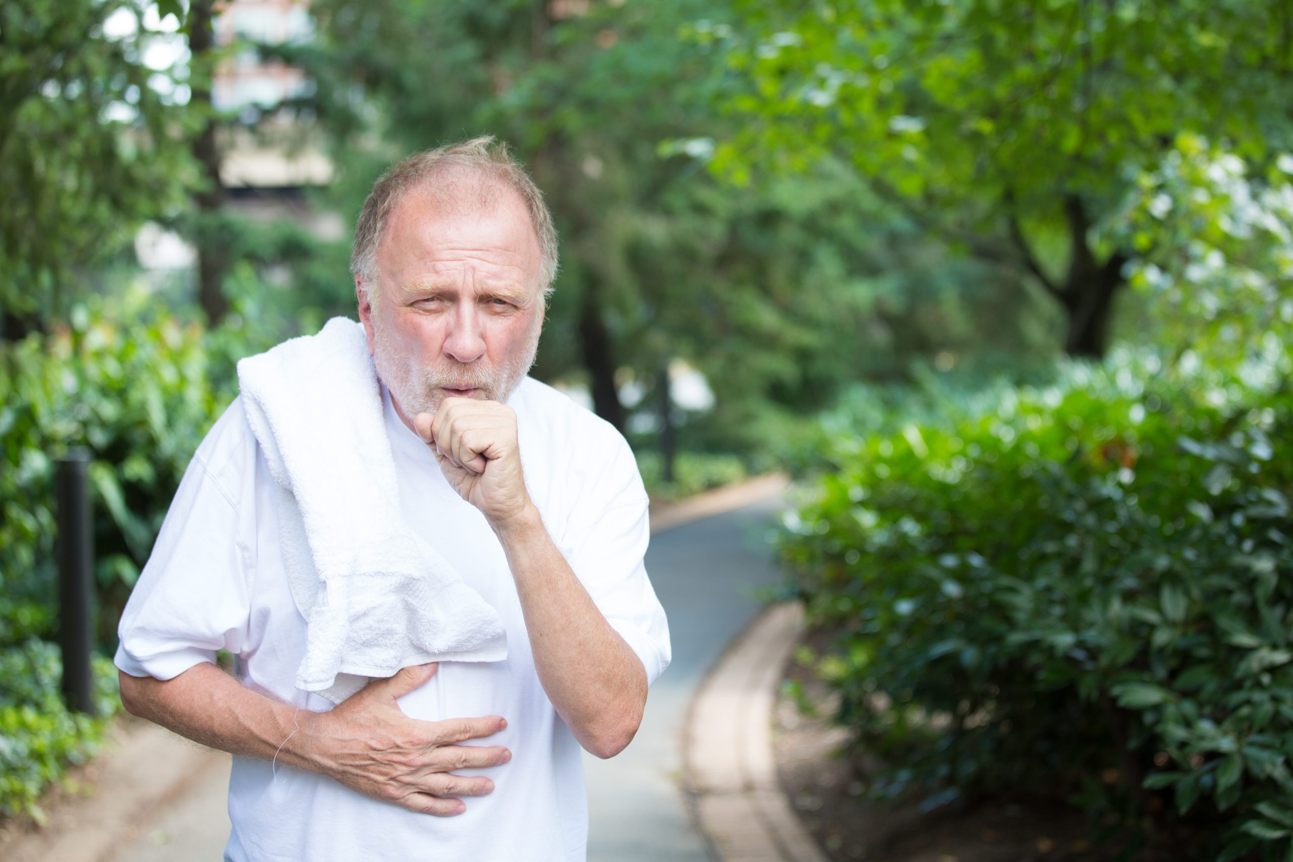 Closeup portrait, old gentleman in white shirt with towel, coughing and holding stomach, isolated green trees and shrubs, outside outdoors background